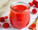 Ein Glas mit leuchtend roter Himbeer-Vinaigrette, umgeben von frischen Himbeeren und einem Holzlöffel mit etwas Vinaigrette.