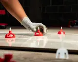 Manos enguantadas colocando niveladores para alicatar sobre pladur. Herramientas rojas y blancas sobre baldosas.