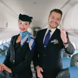 Uśmiechnięta stewardessa i steward w mundurach LOT stoją w samolocie, na tle rzędów siedzeń.