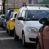 Rząd samochodów, w tym żółty Bolt Taxi, zaparkowany na chodniku.