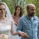 Sposa con velo e bouquet, accompagnata da un uomo in camicia di jeans e una donna sullo sfondo. Scena del **cento domeniche finale**.