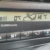 Panel klimatyzacji w samochodzie. Wyświetla 0°C, ikonę nawiewu na szybę i pasażerów, a także ustawienie "HI".