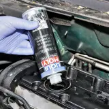 Ręka w niebieskiej rękawiczce wlewa Liqui Moly płukanie silnika do otworu w pokrywie zaworów.