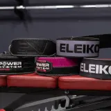 Kilka pasów do dźwigania, w tym marki Eleiko i Power System, leży na czerwonej ławce treningowej.