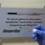 Ręka w niebieskiej rękawiczce trzyma aplikator z bupiwakainą, obok tablica informująca o bezbolesnym znieczuleniu komputerowym SleeperOne.