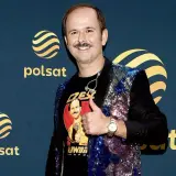 Sławomir Zapała w błyszczącej marynarce i koszulce z własnym wizerunkiem, pokazuje kciuk w górę.