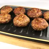 Sieben goldbraune Bouletten, wie nach Omas Rezept, liegen auf einem schwarzen Grillteller, garniert mit Petersilie.