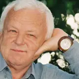 Uśmiechnięty Marian Opaniak, z siwymi włosami i niebieskimi oczami, opiera głowę na dłoni. Zegarek na jego nadgarstku wskazuje godzinę.
