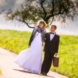 Młoda para w dniu ślubu w maju, idąca polną drogą. Ona w sukni ślubnej i skórzanej kurtce, on w garniturze z walizką.