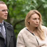 Adam i Ewa obsada: mężczyzna w okularach i płaszczu oraz kobieta w futrzanej kurtce na tle zieleni.