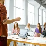 Mężczyzna z mikrofonem prezentuje swój elevator pitch grupie słuchaczy. Na stole laptop z wykresami.