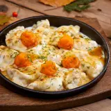 Pierożki z dynią w sosie, posypane koperkiem, podane w żeliwnej patelni na drewnianym blacie. Obok kawałek dyni.