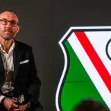 Mężczyzna w okularach, w garniturze, siedzi obok herbu Legii Warszawa. To nowy trener Legii Warszawa.