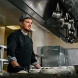 Młody kucharz w czarnym fartuchu i czapce przygotowuje danie w profesjonalnej kuchni.