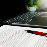 Czerwony długopis leży na dokumencie z poprawkami, obok laptopa. Widać tu różne odmiany cudzysłowu, które autor tekstu musiał poprawić.