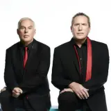 Andy McCluskey i Paul Humphreys z OMD, duet znany z niezapomnianych piosenek, w czarnych garniturach i czerwonych akcentach.