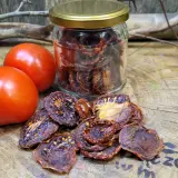 Słoik pełen aromatycznych suszonych pomidorów obok świeżych pomidorów i gałązek na drewnianym blacie.