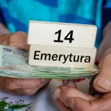 Dłonie trzymają stos banknotów i drewniane klocki z napisem "14 Emerytura".