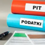 Dwa segregatory: czerwony z napisem "PIT" i niebieski z napisem "PODATKI". Obok formularze podatkowe i długopis. Ulga dla młodych w zasięgu ręki.