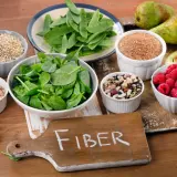 En skärbräda med texten "FIBER" omgiven av skålar med bönor, linser, spenat, hallon, päron, avokado och korn.