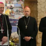 Trzech biskupów w czarnych szatach i z krzyżami na szyjach podczas zjazdu biskupów. Jeden z nich przemawia przy mikrofonie.
