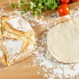 Une recette de pain maison : une miche dorée saupoudrée de farine à côté d'une pâte à pain crue, avec des tomates et du persil en arrière-plan.