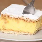 Kawałek kremowej napoleonki z cukrem pudrem, jak z przepisu babci. Widelec zagłębia się w puszysty krem.