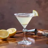 Klasyczny martini z cytryną i solą na kieliszku. W tle cytryny, oliwki i akcesoria barmańskie. Poznaj różne martini rodzaje.