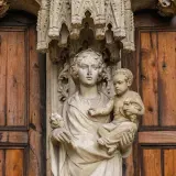 Kamienna rzeźba Matki Boskiej z Dzieciątkiem na tle drewnianych drzwi, symbolizująca teocentryzm i boską obecność w codzienności.