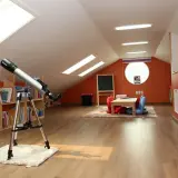 Na bardzo niskim i małym poddaszu stoi teleskop, obok regały z książkami i kącik do zabawy z tablicą.
