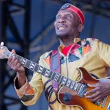 Ein verstorbener Musiker, eine Legende, spielt Gitarre auf der Bühne. Sein buntes Outfit und sein Ausdruck zeugen von seiner Leidenschaft.