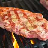Un steak de basse côte, assaisonné, grésille sur un barbecue. Les flammes dansent sous la grille, promettant une cuisson parfaite.
