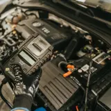Mechanik w rękawiczkach sprawdza stan akumulatora za pomocą testera, który może również diagnozować czujnik halla w samochodzie.