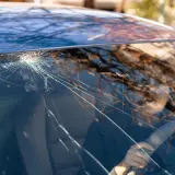 Un parabrezza rotto mostra una ragnatela di crepe che si irradiano da un punto d'impatto. Riflessi di alberi e una persona al volante sono visibili attraverso il vetro danneggiato.