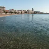 El mar Mediterráneo baña la arena dorada de la playa Levante en Santa Pola, con edificios modernos y palmeras al fondo.
