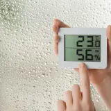 Manos sostienen un higrómetro digital que muestra 23.0°C y 56% de humedad, mientras gotas de lluvia cubren el cristal.