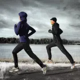 Zwei Läuferinnen in warmer Winterkleidung joggen am Fluss entlang. Die eine trägt einen blauen Hoodie und Hijab, die andere eine schwarze Jacke und orangefarbenen Schal.