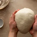 Mani che impastano un panetto soffice, ingrediente chiave per imparare come fare il lievito madre. Farina e ciotolina a lato.