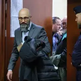 Mężczyzna w garniturze z książką i kurtką, obok policjant i inni mężczyźni w pomieszczeniu.