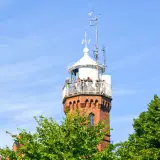 Widok na ceglaną latarnię morską w Ustce z tarasem widokowym, na którym stoją ludzie. W tle błękitne niebo i zielone drzewa.