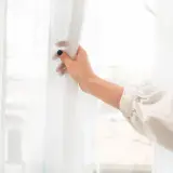 Dłoń odsłaniająca białą firankę. Dowiedz się, jak wybielić firanki w pralce, by znów lśniły bielą.