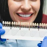 Uśmiechnięta kobieta z idealnymi zębami, dentysta pokazuje paletę kolorów do wyboru najtańszych licówek na zęby.