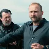 Dwóch mężczyzn na dachu, jeden w skórzanej kurtce, drugi w czarnej koszuli. Scena z jednego z juliusz machulski filmów.