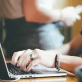 Dłonie piszą na laptopie, przygotowując księgowanie alior. W tle widać inne osoby i roślinę.
