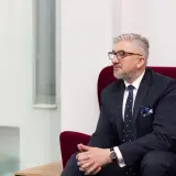 Mężczyzna w garniturze siedzi w czerwonym fotelu, patrząc w bok.