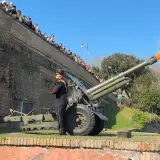 Il cannone Gianicolo spara una palla di fumo arancione e gialla. Un soldato in uniforme osserva, mentre una folla guarda dall'alto.