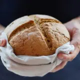 Dłonie trzymają okrągły bochenek chleba, owinięty w lnianą ściereczkę. Panie dobry jak chleb, symbolizuje świeżość i tradycję.