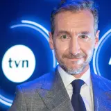 Uśmiechnięty mężczyzna w garniturze na tle logo TVN. Jeden z dziennikarzy Wiadomości TVN, gotowy do przekazania najnowszych informacji.
