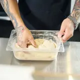 Ręce z tatuażami zagniatają ciasto na pizzę w pojemniku. Czy ciasto na pizzę można mrozić? Tak, to ciasto jest przygotowane do mrożenia.