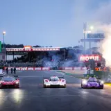 Tres coches de carreras compiten en Le Mans, con fuegos artificiales iluminando la pista. El horario de Le Mans se acerca.